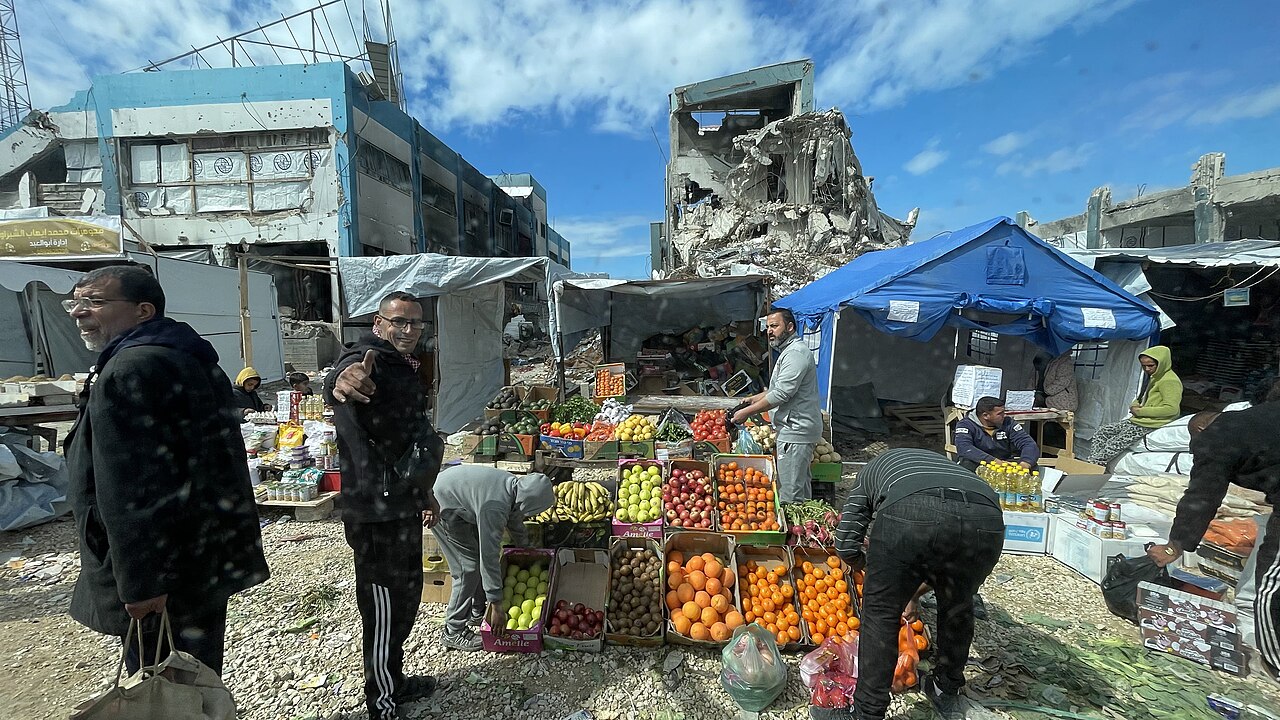 Displaced families living in tents in Gaza, a fruit market set up among the tents and destroyed buildings behind